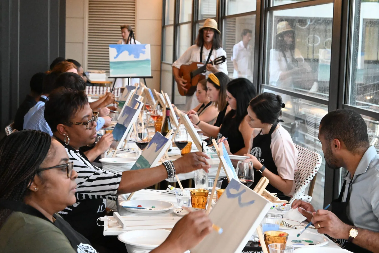 Group painting along with the instructor's example canvas while a guitarist plays